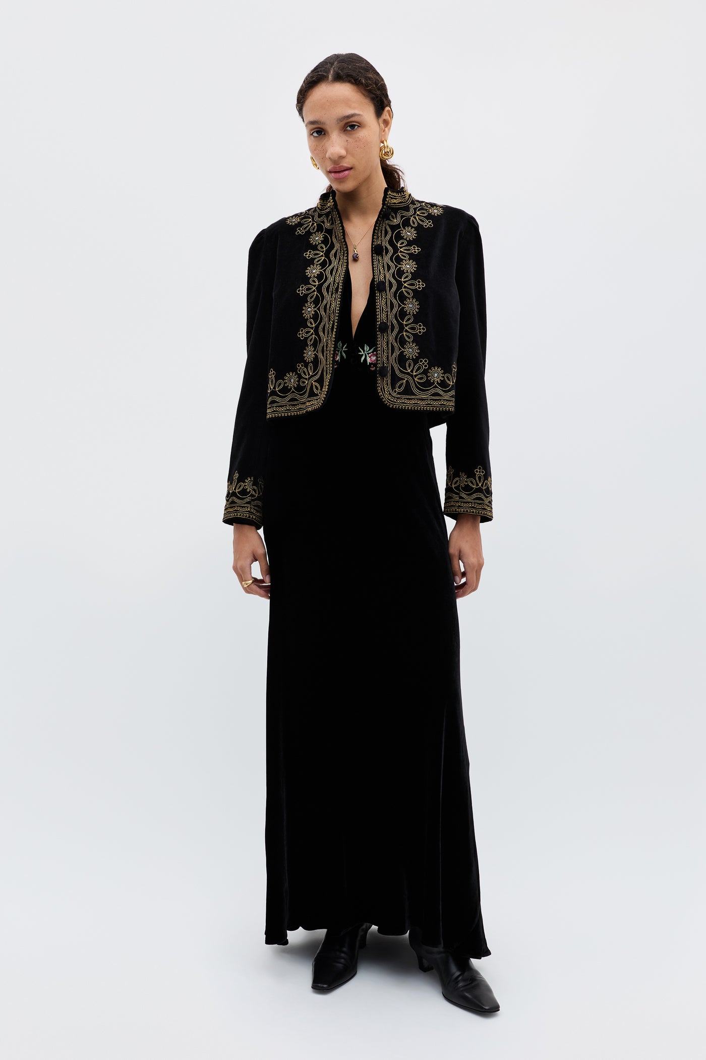 A woman wears the Emory - Blossom Embroidery Pink cropped jacket over a 1930s-inspired long black dress with black boots, standing against a plain white background and looking directly at the camera with a neutral expression.