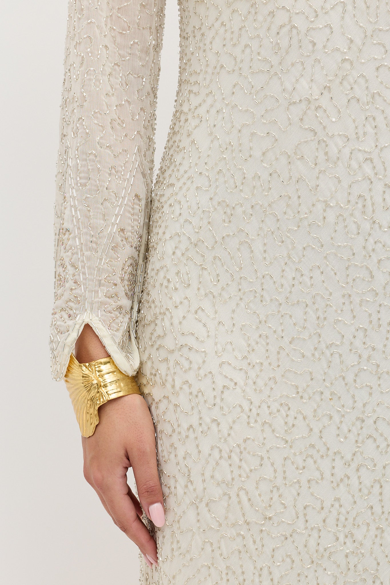 Wearing the Alondra - Florence Embellishment Pearl cream dress with pearl details, a person rests their hand by their side, showing light pink nails and a wide gold cuff bracelet featuring a butterfly design.