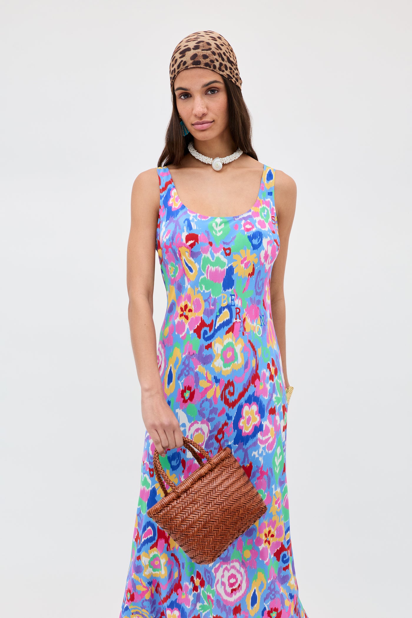 A woman in the Bondi - Ikat Bloom Lilac floral dress, with a leopard print headscarf, pearl choker, and woven brown handbag, stands against a plain white background.