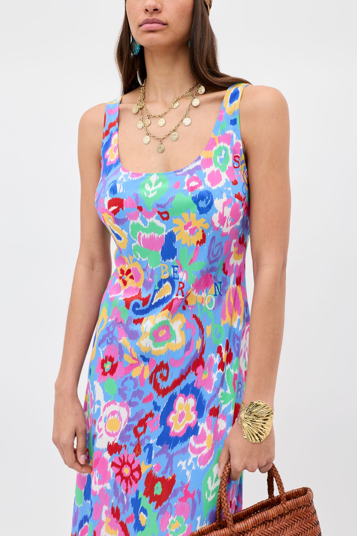 A woman in the Bondi - Ikat Bloom Lilac sleeveless floral dress with a scoop neckline, gold coin necklaces, a gold leaf bracelet, and holding a woven brown bag. She has long, straight hair.