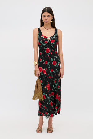 A woman wears the Bondi - Claudia Rose Black, a sleeveless ankle-length black slip dress with a red rose floral print, styled with gold jewelry, nude heels, and a gold tasseled handbag against a plain white background.