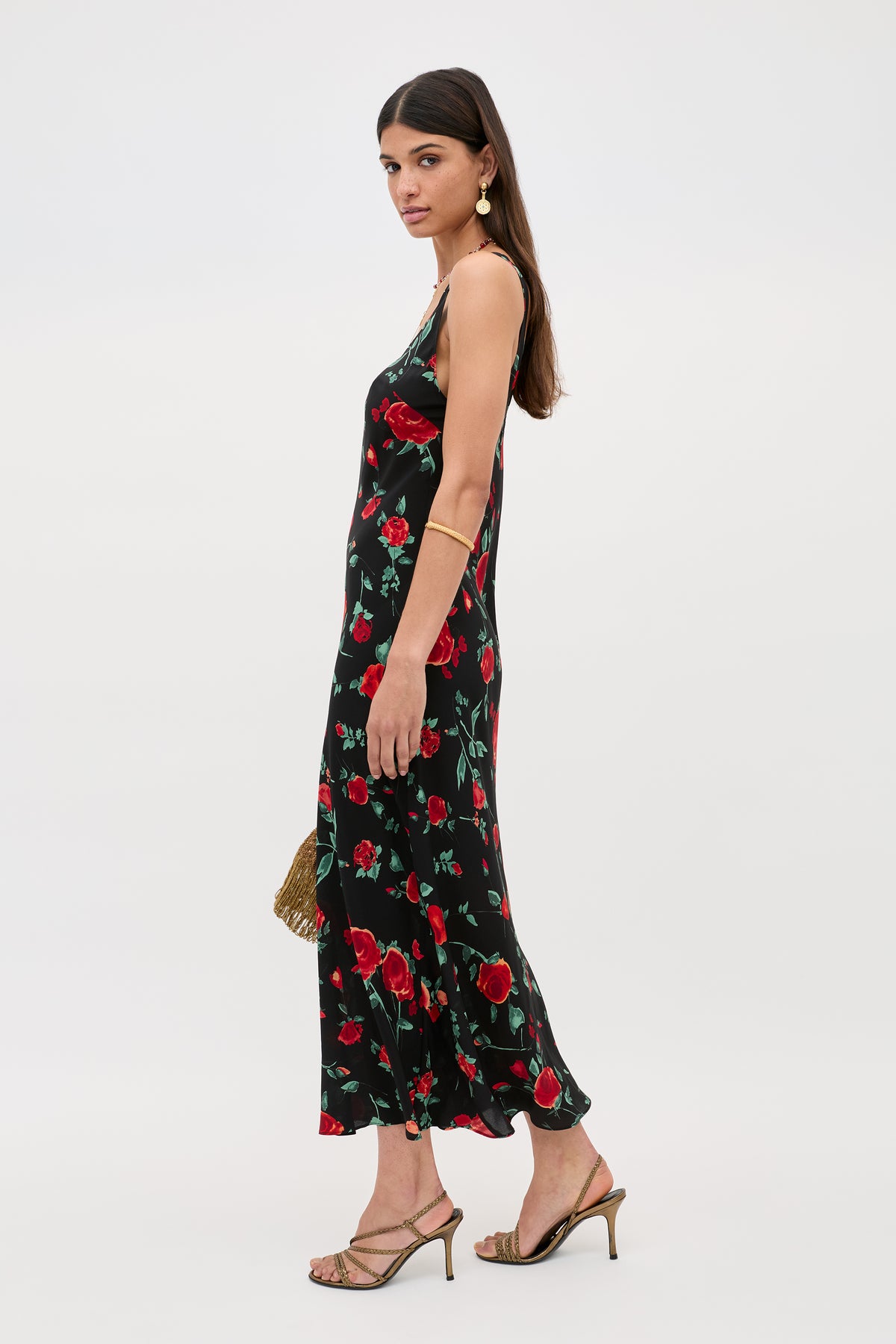 Woman in a sleeveless Bondi - Claudia Rose Black midi dress with a red rose print, gold drop earrings, and gold strappy heels, standing in profile against a plain light background.