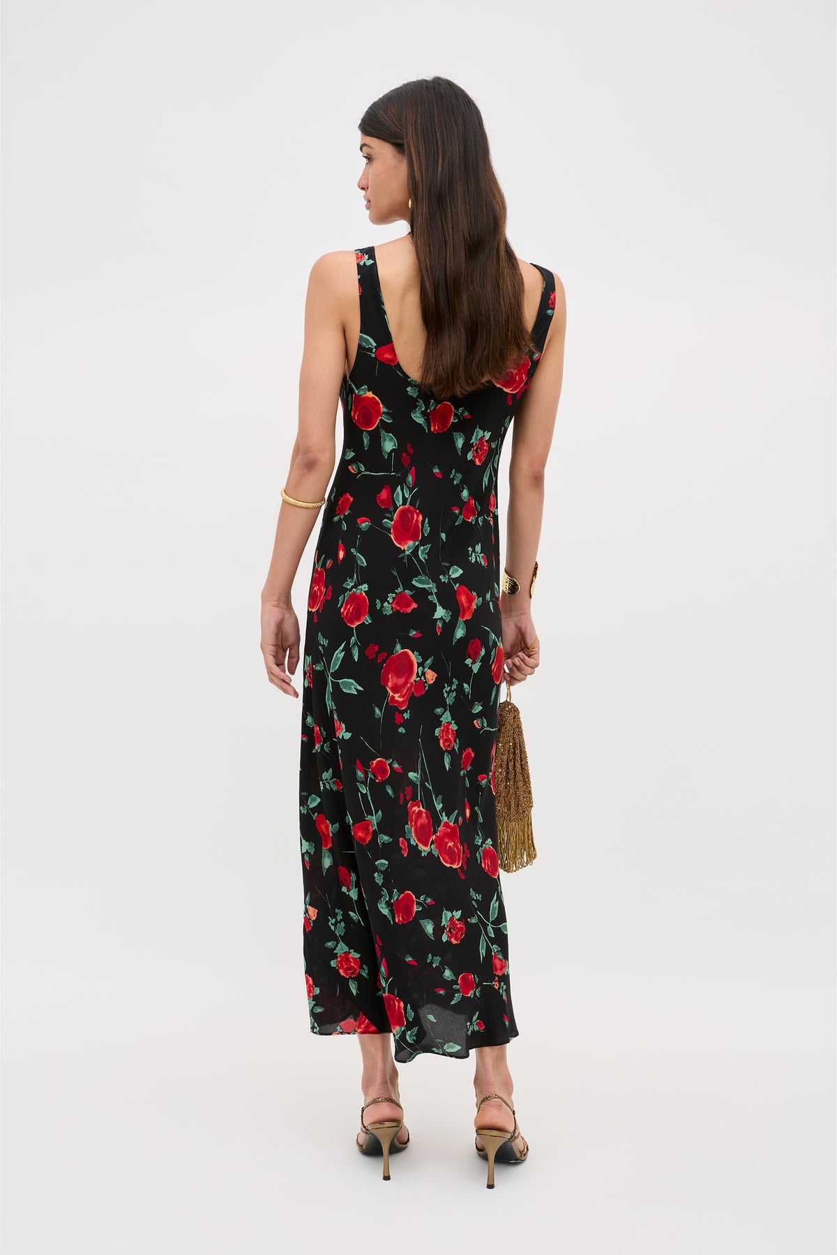 A woman faces away, wearing the Bondi - Claudia Rose Black sleeveless slip dress with red rose pattern and bias silhouette. She holds a gold fringe handbag, wears black heels, and her long brown hair falls down her back against a plain light background.