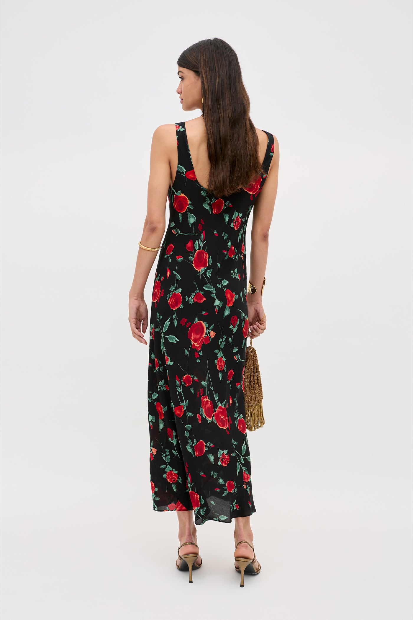 A woman faces away, wearing the Bondi - Claudia Rose Black sleeveless slip dress with red rose pattern and bias silhouette. She holds a gold fringe handbag, wears black heels, and her long brown hair falls down her back against a plain light background.