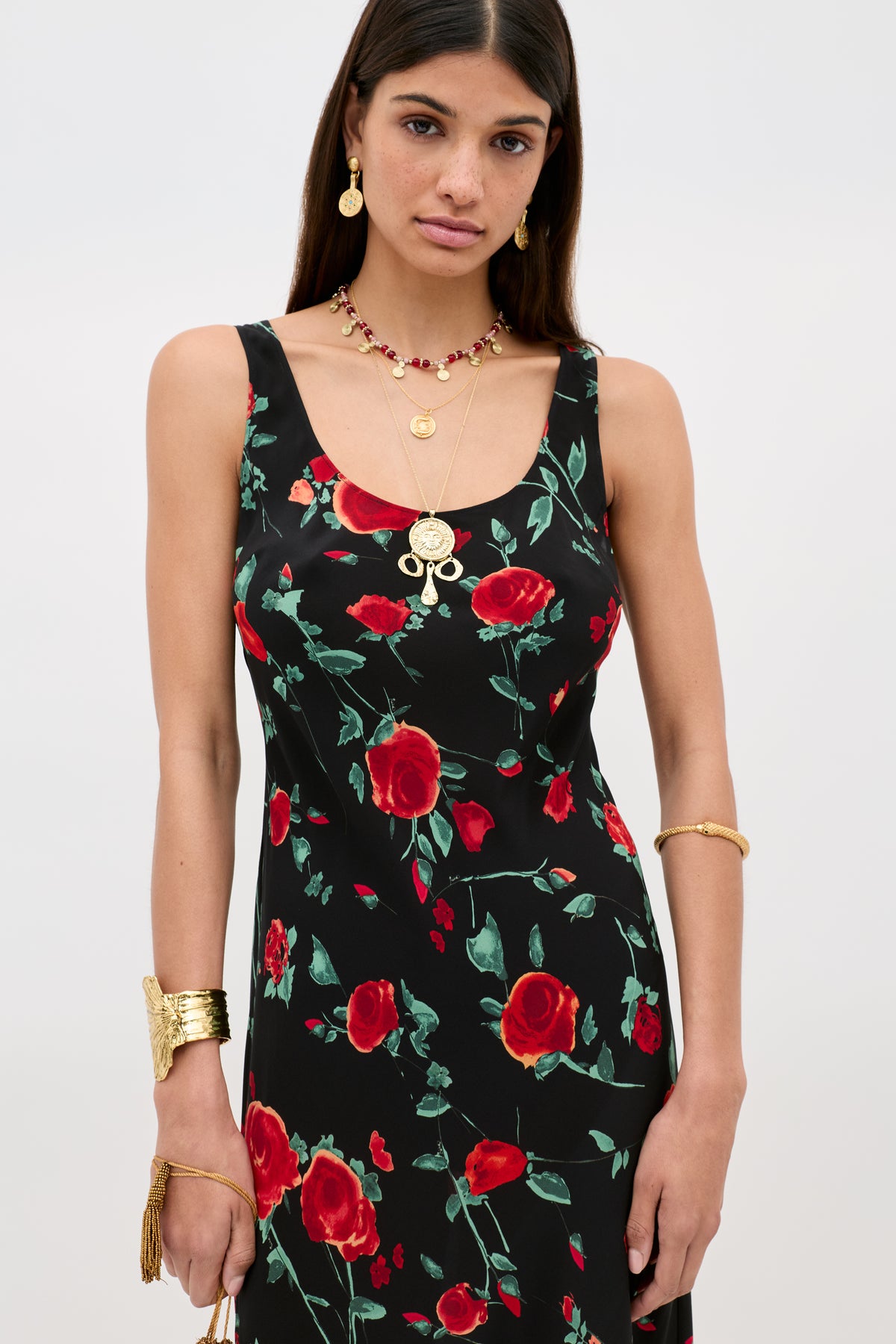 A woman in the Bondi - Claudia Rose Black sleeveless slip dress with a red rose print faces the camera. She pairs it with layered gold necklaces, earrings, a bracelet, and a cuff. Her long hair is straight.