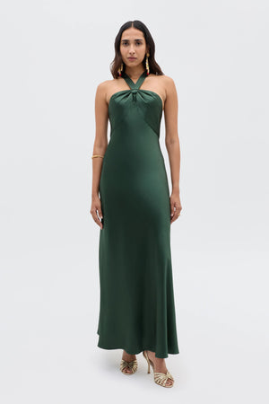 A woman wears the Losanna - Forest Green dress, a long satin halter style with a low back. She pairs it with strappy gold heels, gold earrings, and a bracelet. Her long dark hair is styled loosely—a perfect choice for elegant evenings.