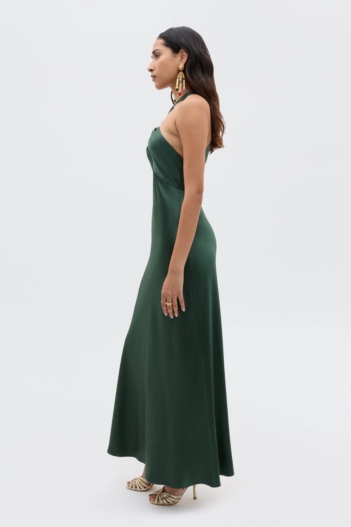A woman stands in profile against a white background wearing the Losanna - Forest Green sleeveless, floor-length satin dress with gold high-heeled sandals and large statement earrings.