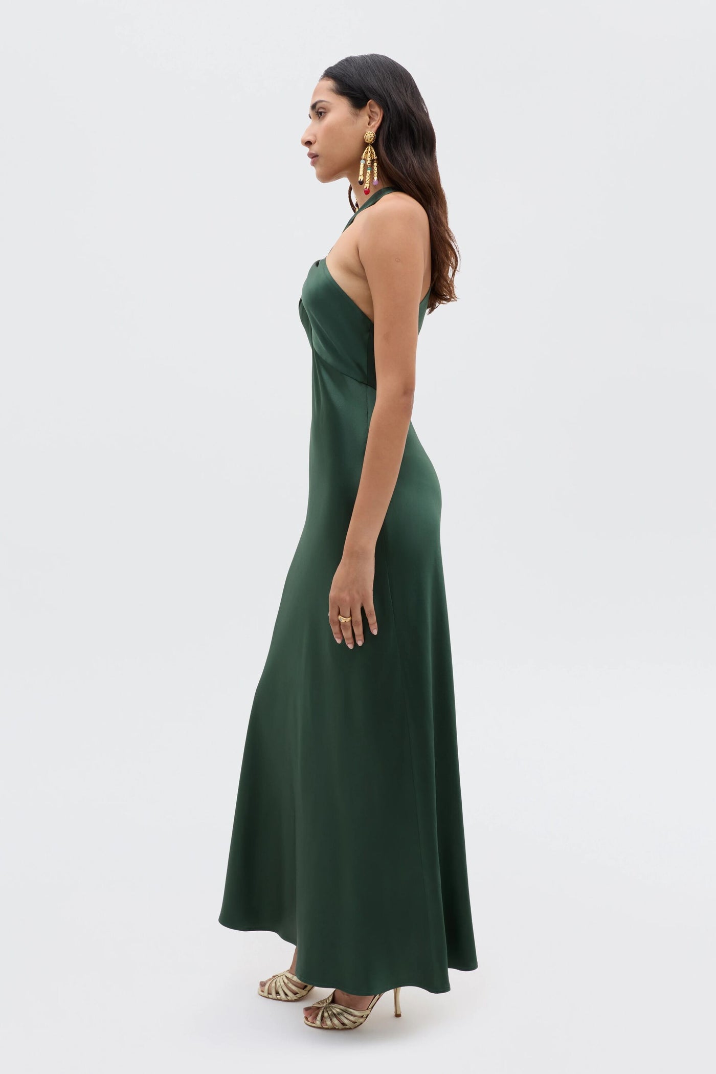 A woman stands in profile against a white background wearing the Losanna - Forest Green sleeveless, floor-length satin dress with gold high-heeled sandals and large statement earrings.