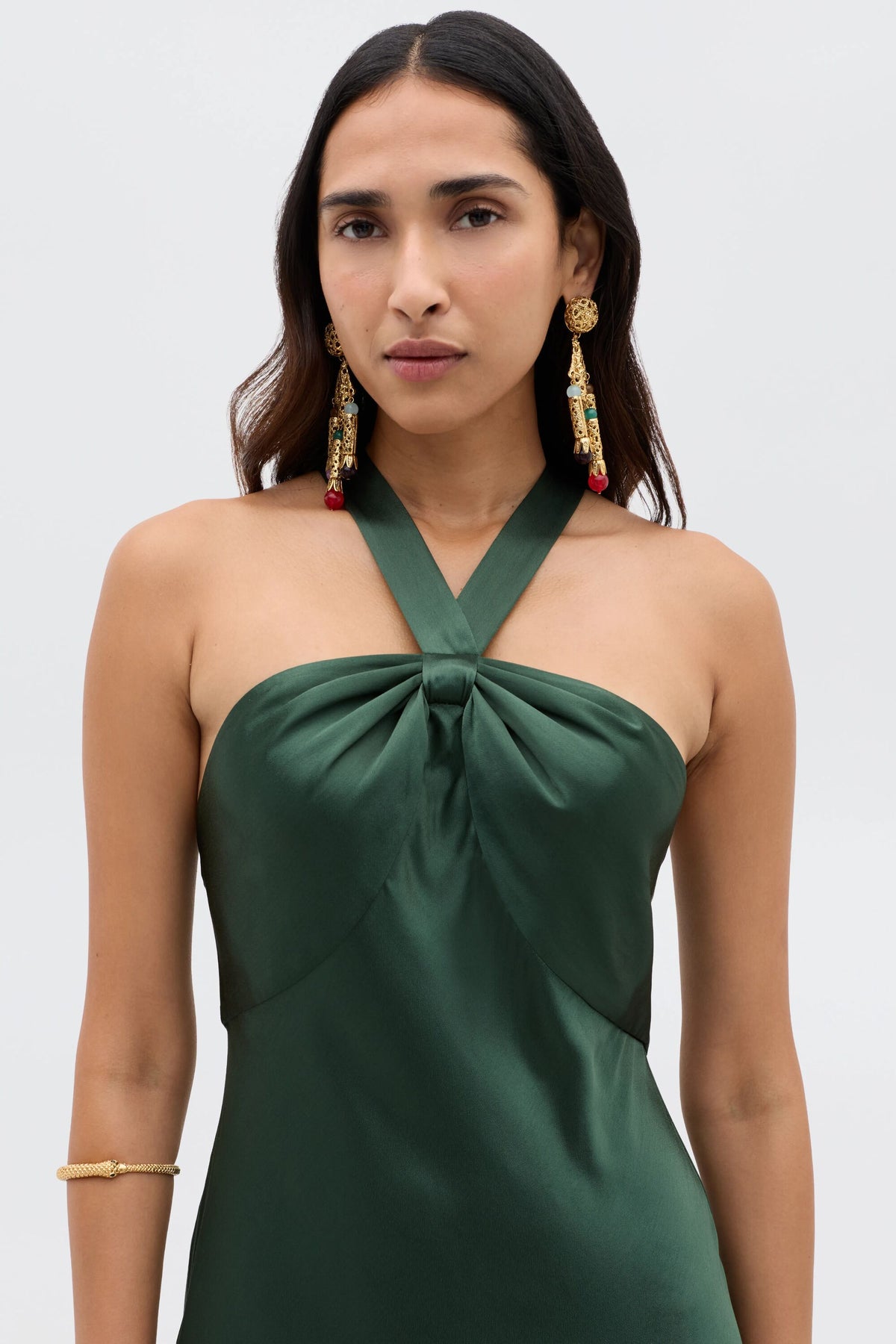 A woman with long dark hair wears the Losanna - Forest Green satin halter-neck evening dress, gold earrings with red accents, and a gold bracelet, standing against a plain light gray background.