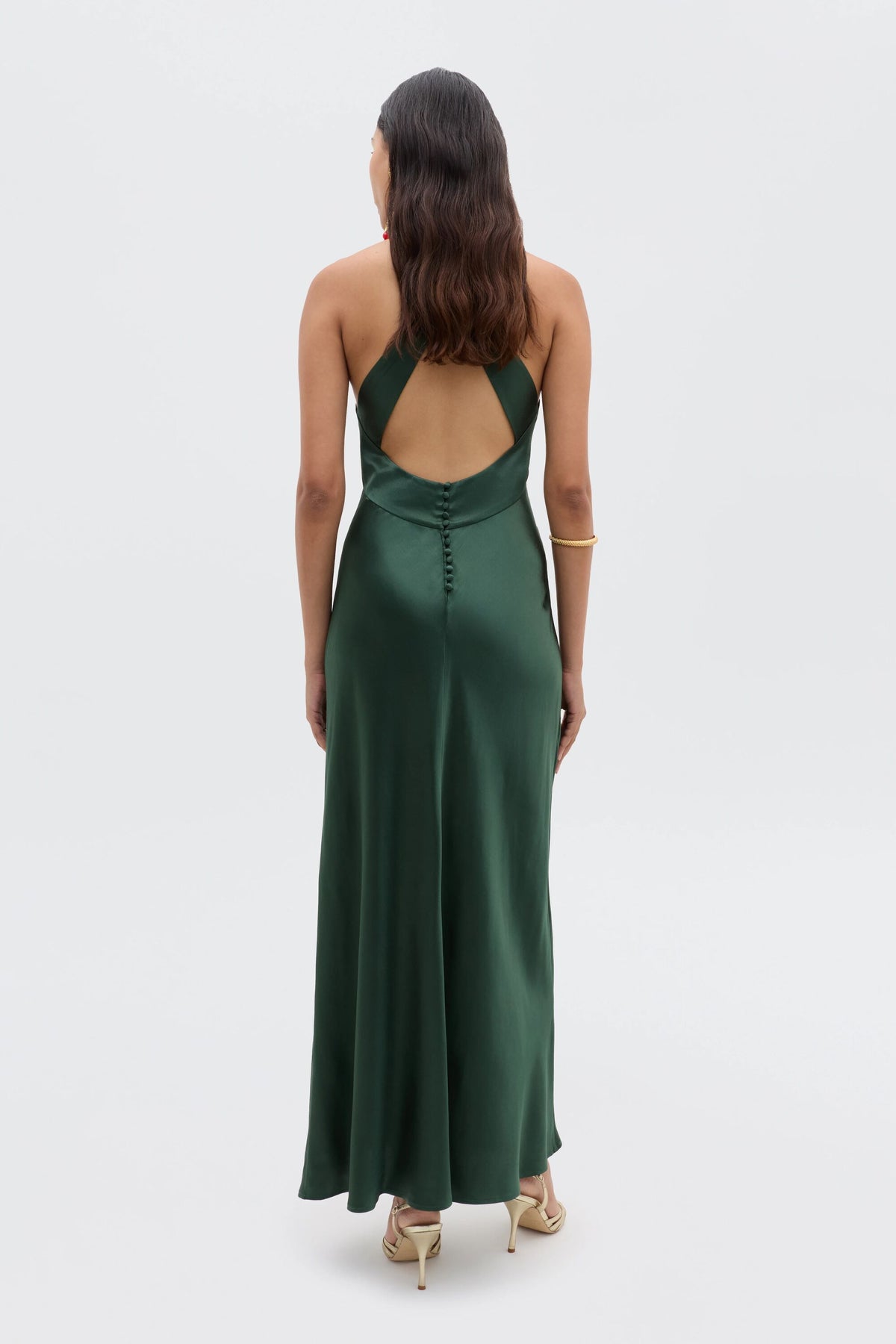 A woman faces away, highlighting the low back design of her long Losanna - Forest Green gown. She wears her hair in waves, pairs the dress with a gold bracelet and light-colored heels for an elegant evening look.