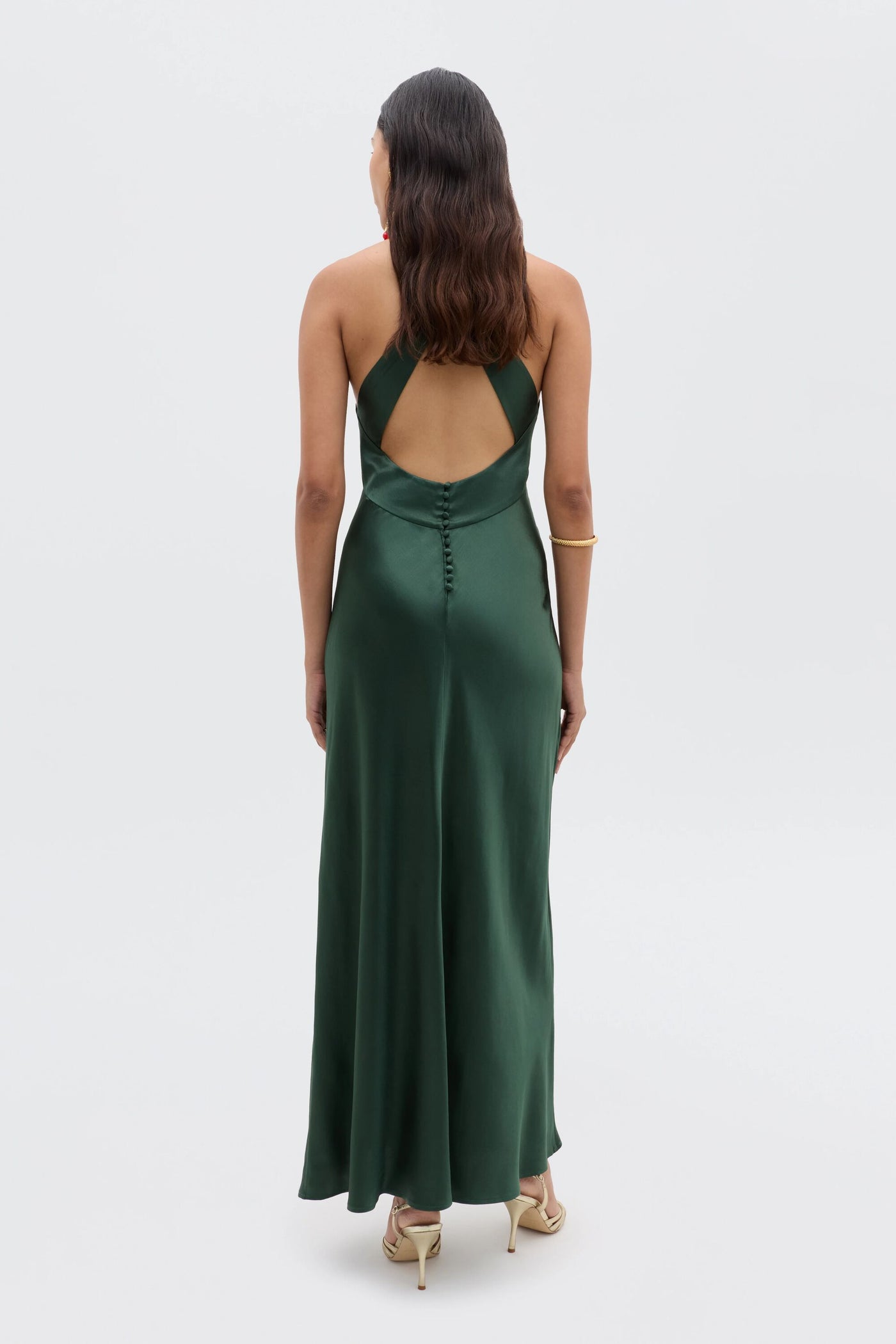 A woman faces away, highlighting the low back design of her long Losanna - Forest Green gown. She wears her hair in waves, pairs the dress with a gold bracelet and light-colored heels for an elegant evening look.