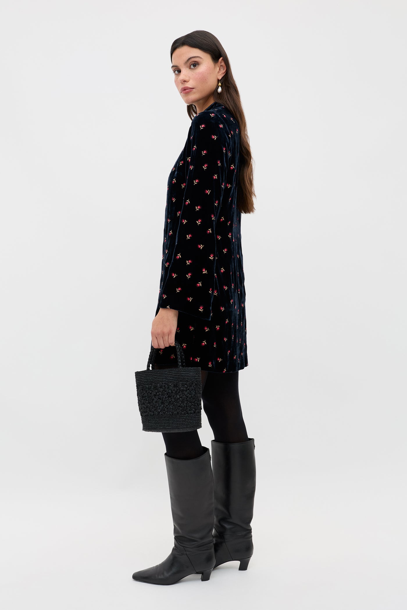 A woman with long brown hair stands sideways against a white background, wearing the Calla - Dandy Embroidery Deep Navy dress, black tights, tall black boots, and holding a textured black handbag.