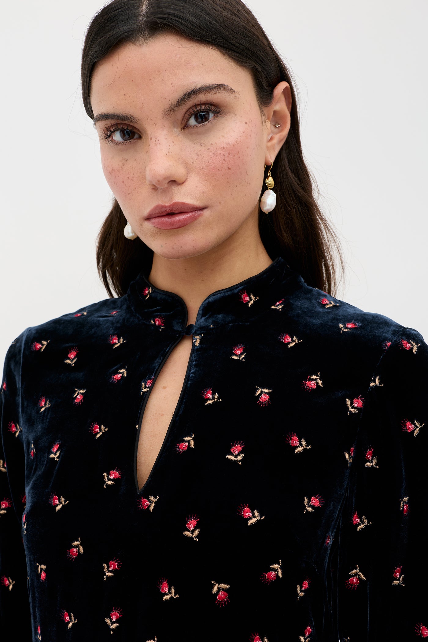 A woman with long dark hair, wearing pearl drop earrings and the Calla - Dandy Embroidery Deep Navy velvet party dress with a keyhole neckline, looks directly at the camera against a plain light background.