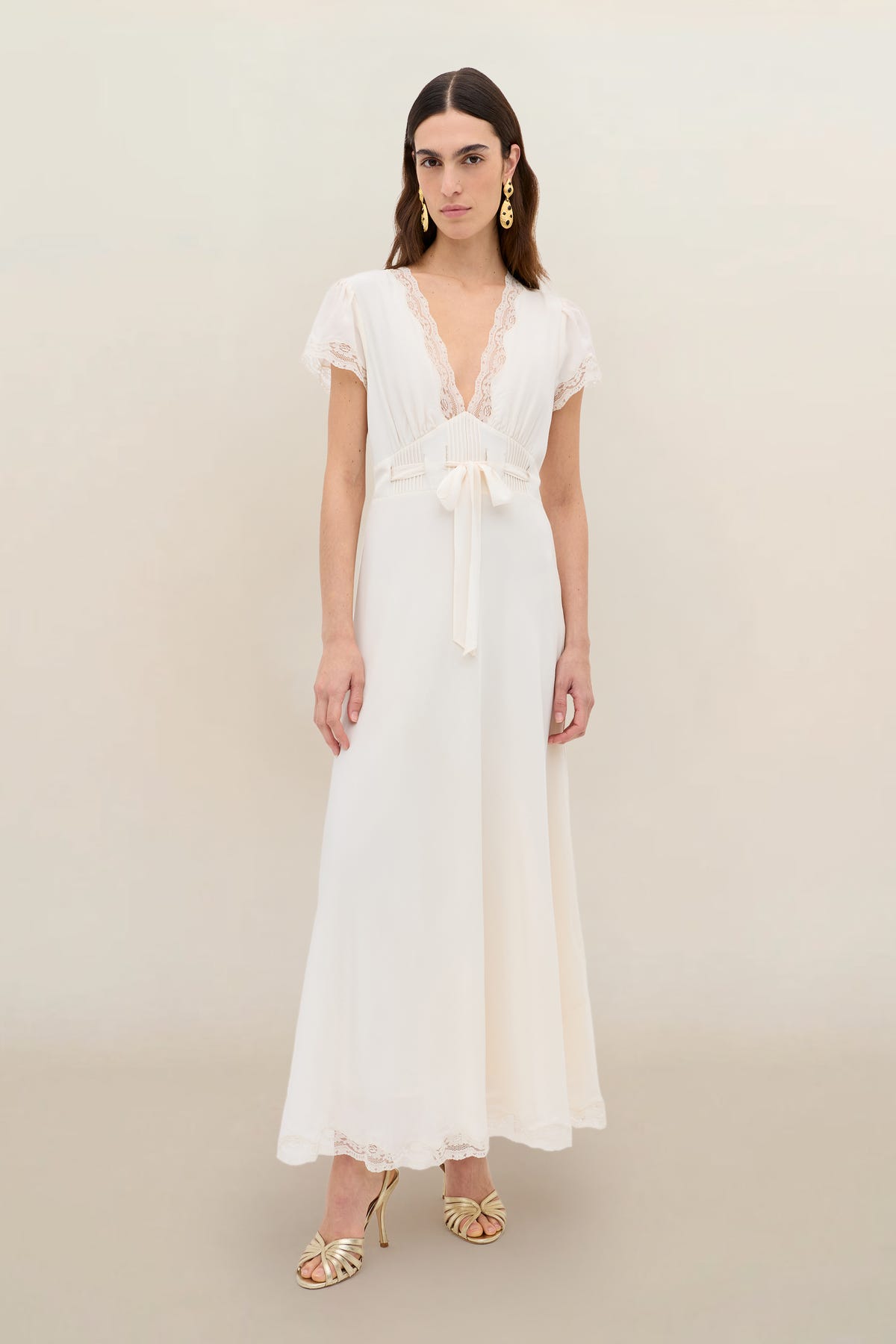 Wearing the Marlice - Pearl, a long, vintage-inspired white dress with lace trim and a deep V-neckline, she stands against a light background. Her look is finished with gold earrings, open-toe heels, and loose, center-parted hair.