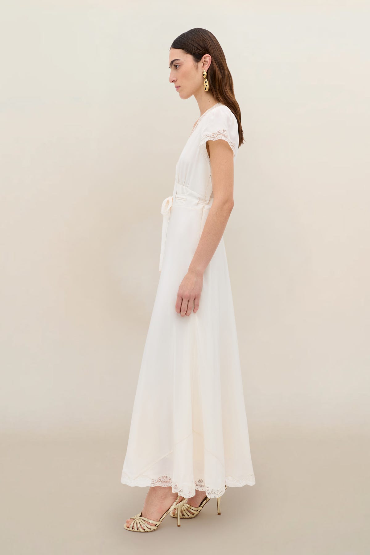 A woman stands in profile against a neutral background, wearing the Marlice - Pearl: a vintage-inspired, ankle-length ivory dress with lace trim, satin bow detail, gold earrings, and strappy heels.