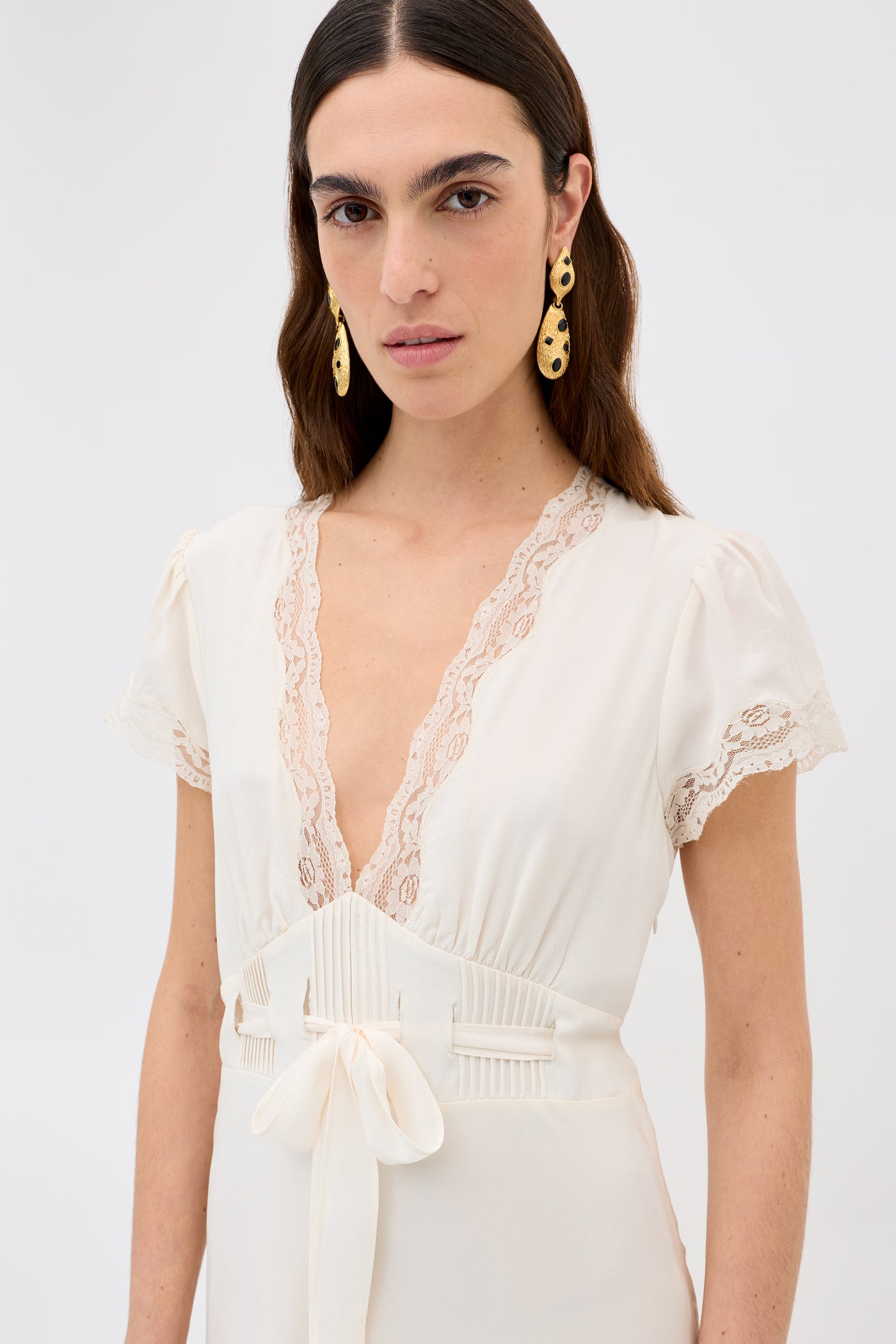 A woman with long dark hair wears gold earrings and the Marlice - Pearl dress, a cream vintage-inspired piece with lace trim, short sleeves, a deep V-neckline, and a satin bow waist. She stands against a plain light background.