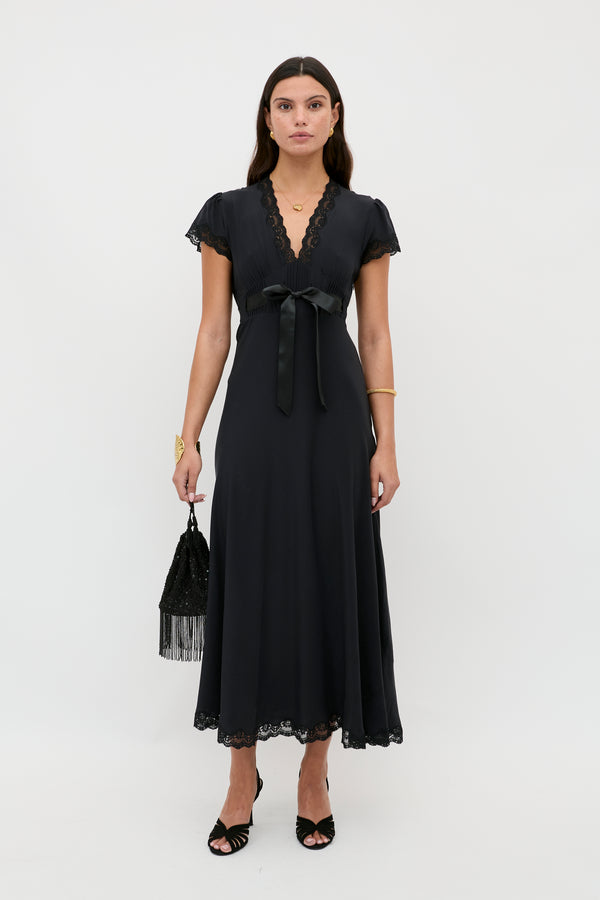 Wearing the Marlice - Black dress—a short-sleeved, V-neck, ankle-length design with lace trim and a satin waist bow—a woman holds a black fringe handbag and wears black sandals, exemplifying classic elegance against a white background.