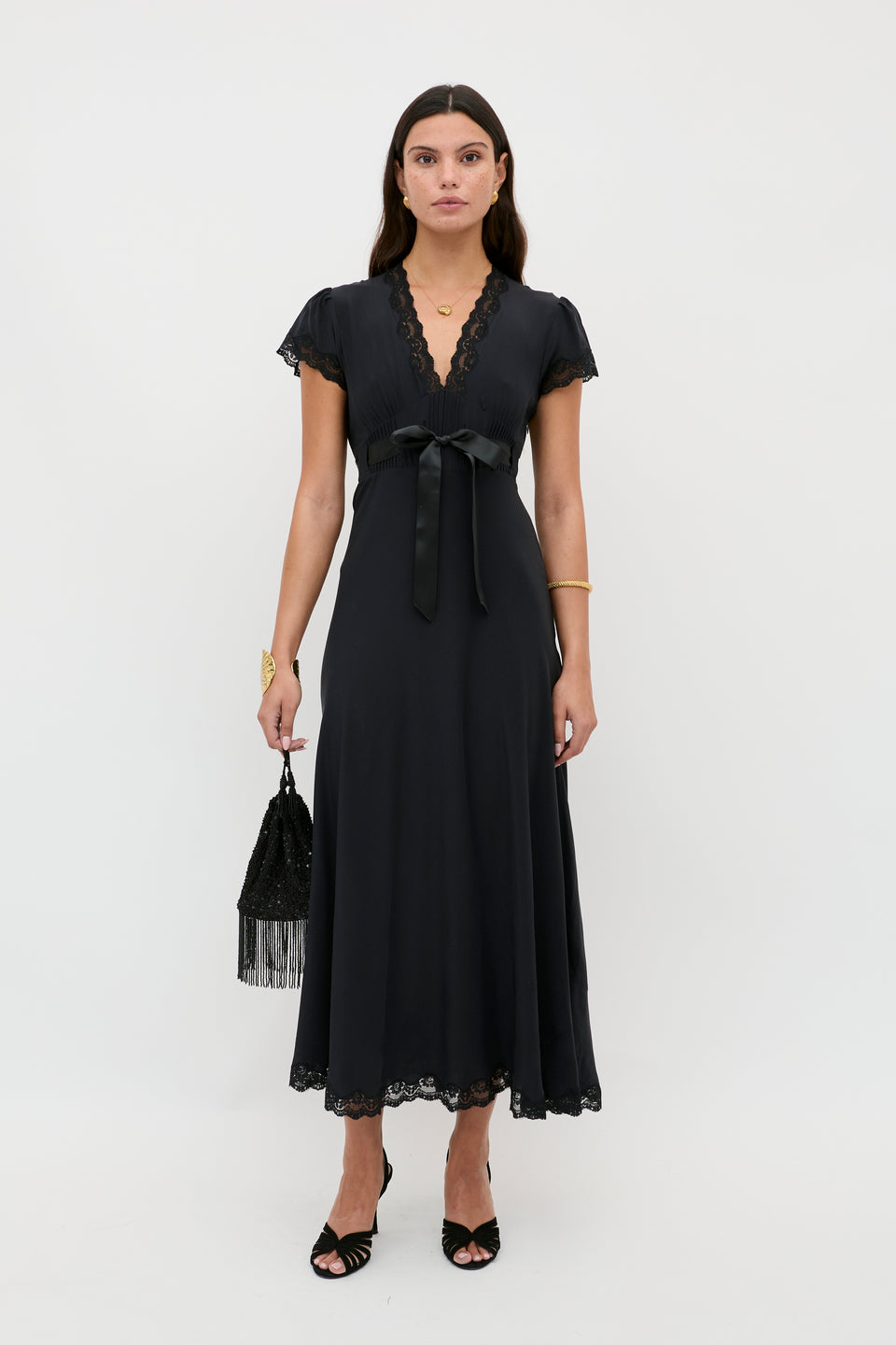 Wearing the Marlice - Black dress—a short-sleeved, V-neck, ankle-length design with lace trim and a satin waist bow—a woman holds a black fringe handbag and wears black sandals, exemplifying classic elegance against a white background.