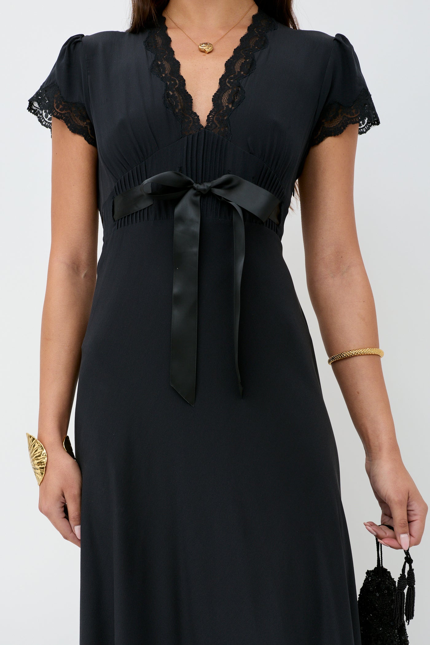 Woman wearing the Marlice - Black dress, featuring lace trim, short sleeves, a V-neckline, and a satin waist bow. She accessorizes with gold jewelry and carries a small black bag—ideal for lovers of chic black dresses.