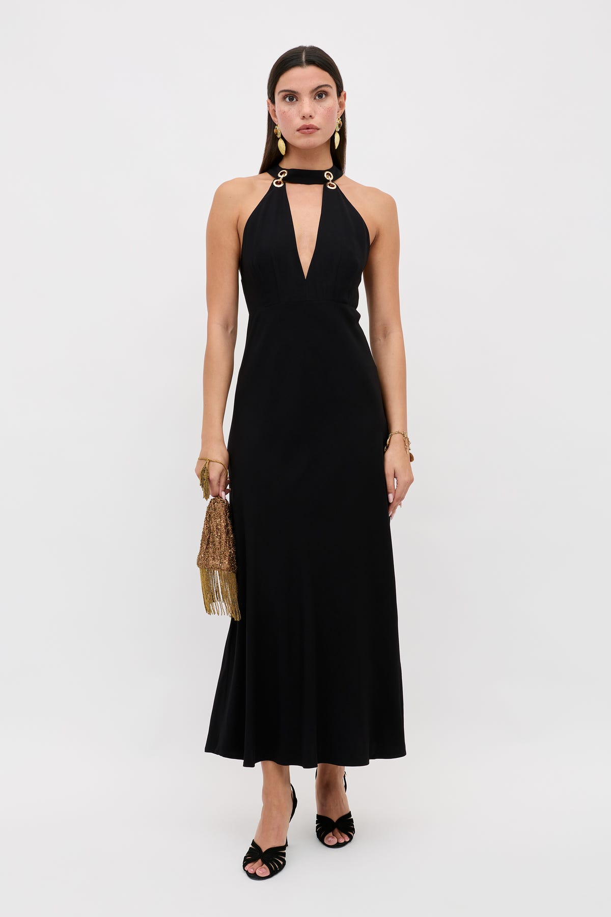 A woman poses against a white background wearing the Eleanora - Black sleeveless, ankle-length dress featuring a deep V neckline and gold ring shoulder details, accessorized with a gold fringe handbag and black open-toe heels.