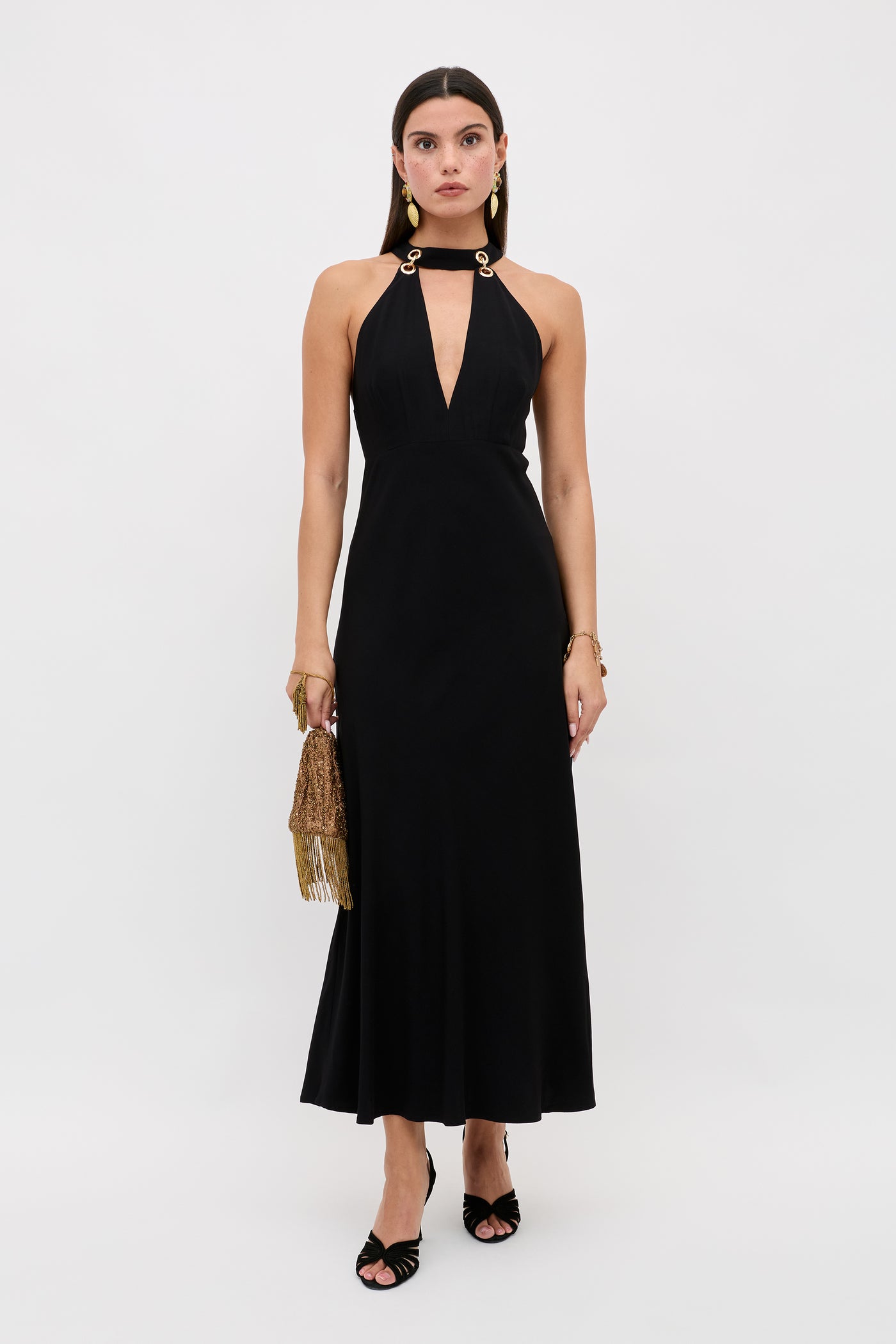 A woman poses against a white background wearing the Eleanora - Black sleeveless, ankle-length dress featuring a deep V neckline and gold ring shoulder details, accessorized with a gold fringe handbag and black open-toe heels.