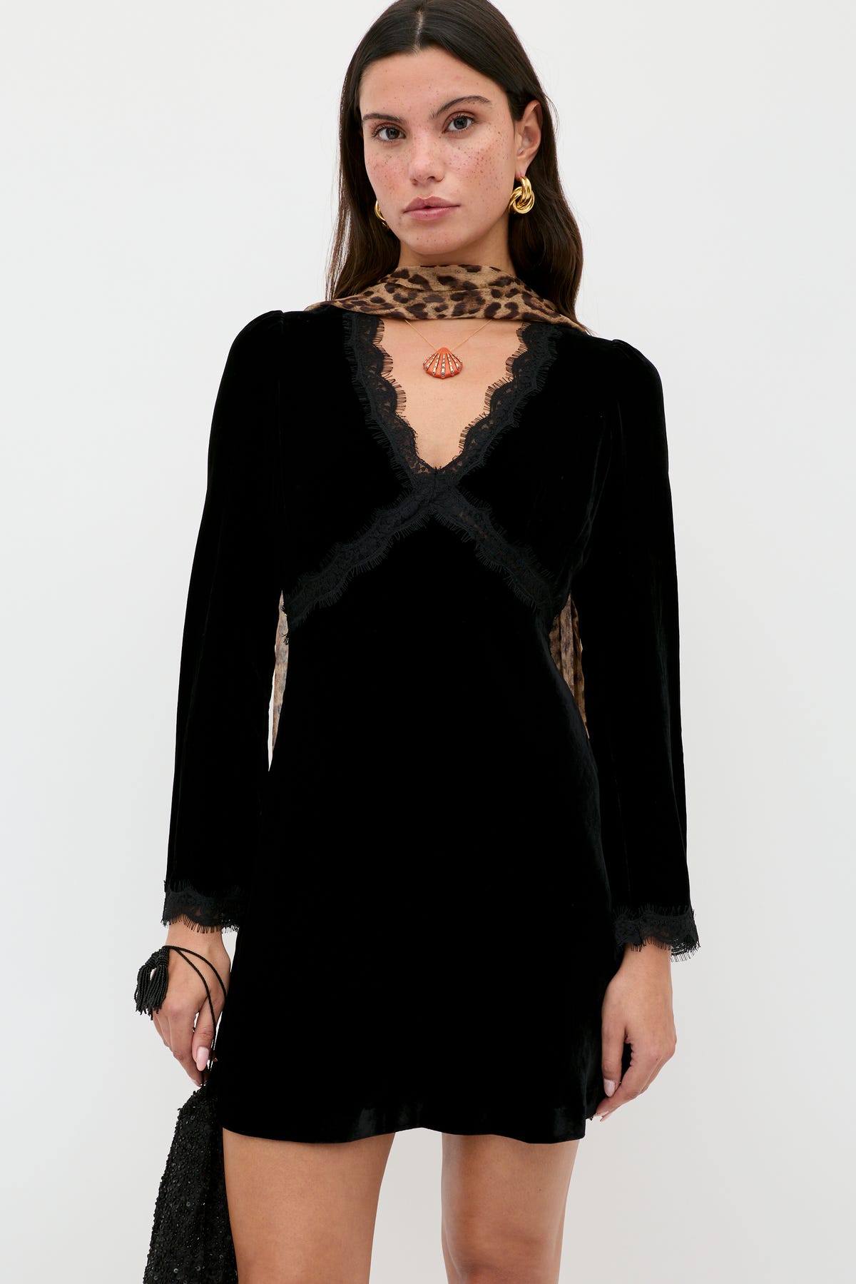 A woman stands against a white background wearing the RIXO Macie - Black dress with lace trim and long sleeves, accessorized with gold earrings, a leopard print scarf, and holding a black purse.