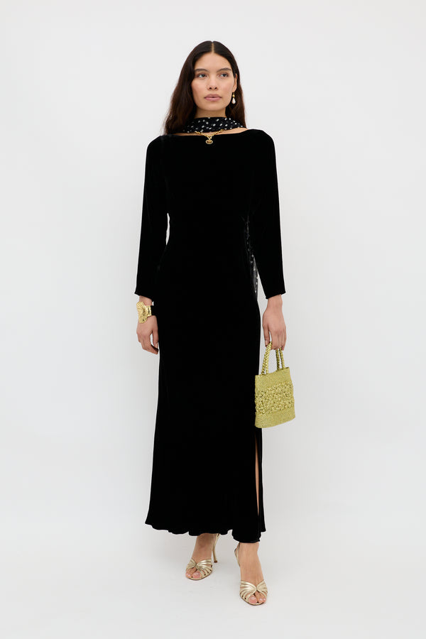 A woman in an elegant long-sleeve evening dress with a slit, gold sandals, a gold bracelet, and a yellow woven handbag stands against a white background, wearing the Devina - Black scarf with white polka dots around her neck.