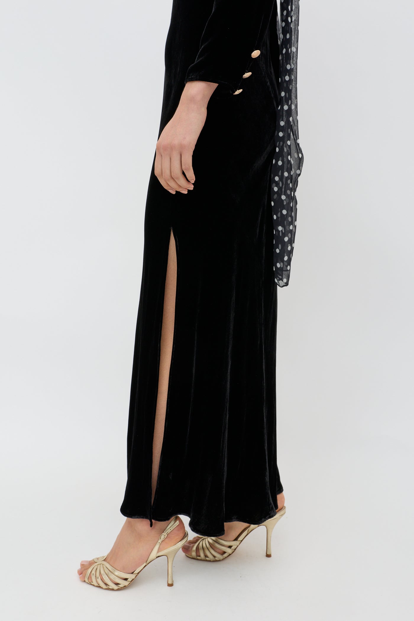 A person wearing the Devina - Black, a long black velvet evening gown with a side slit, gold heels, and a sheer black polka dot scarf stands against a plain white background.