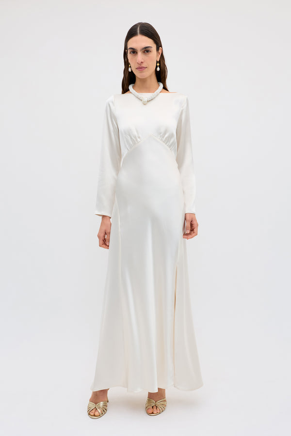 A woman stands against a white background wearing the Devina - Pearl, a long-sleeved, floor-length ivory satin dress with a high waist and subtle slit, styled with a pearl necklace, drop earrings, and gold open-toe heels.