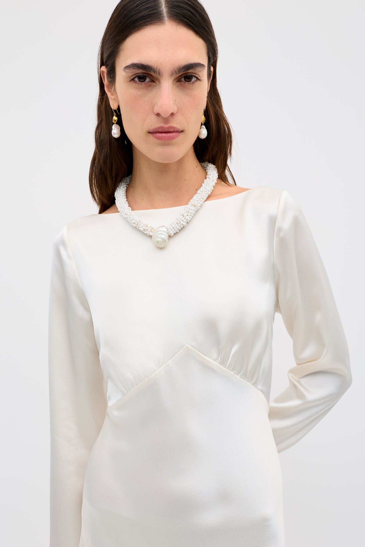 A woman with long dark hair wears the Devina - Pearl dress, paired with pearl earrings and a chunky pearl necklace, against a plain light background.