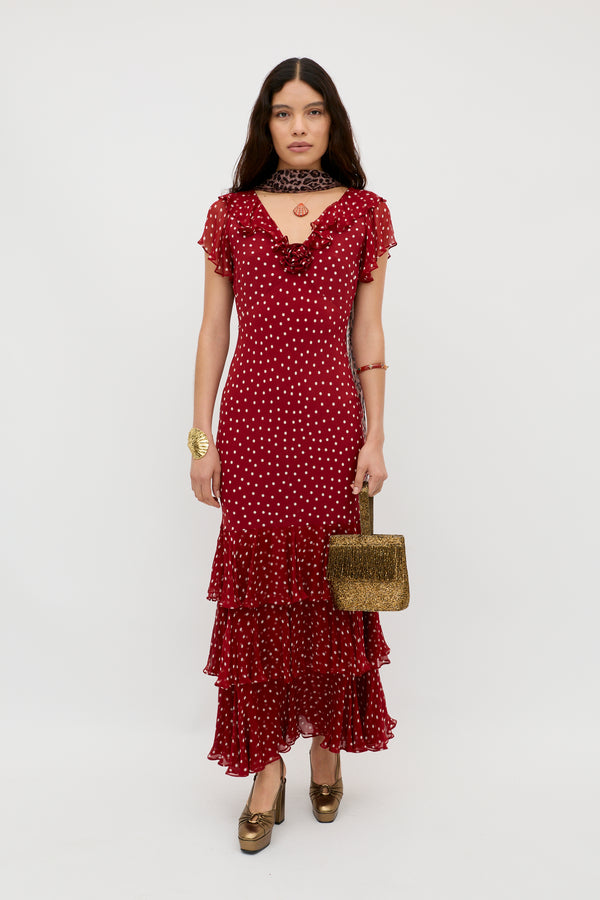 A woman poses against a plain background wearing the Amelle - Layla Dot Red dress, featuring short sleeves, polka dots, and tiered ruffles. She pairs it with gold heels, a choker, bracelet, and a textured gold handbag.