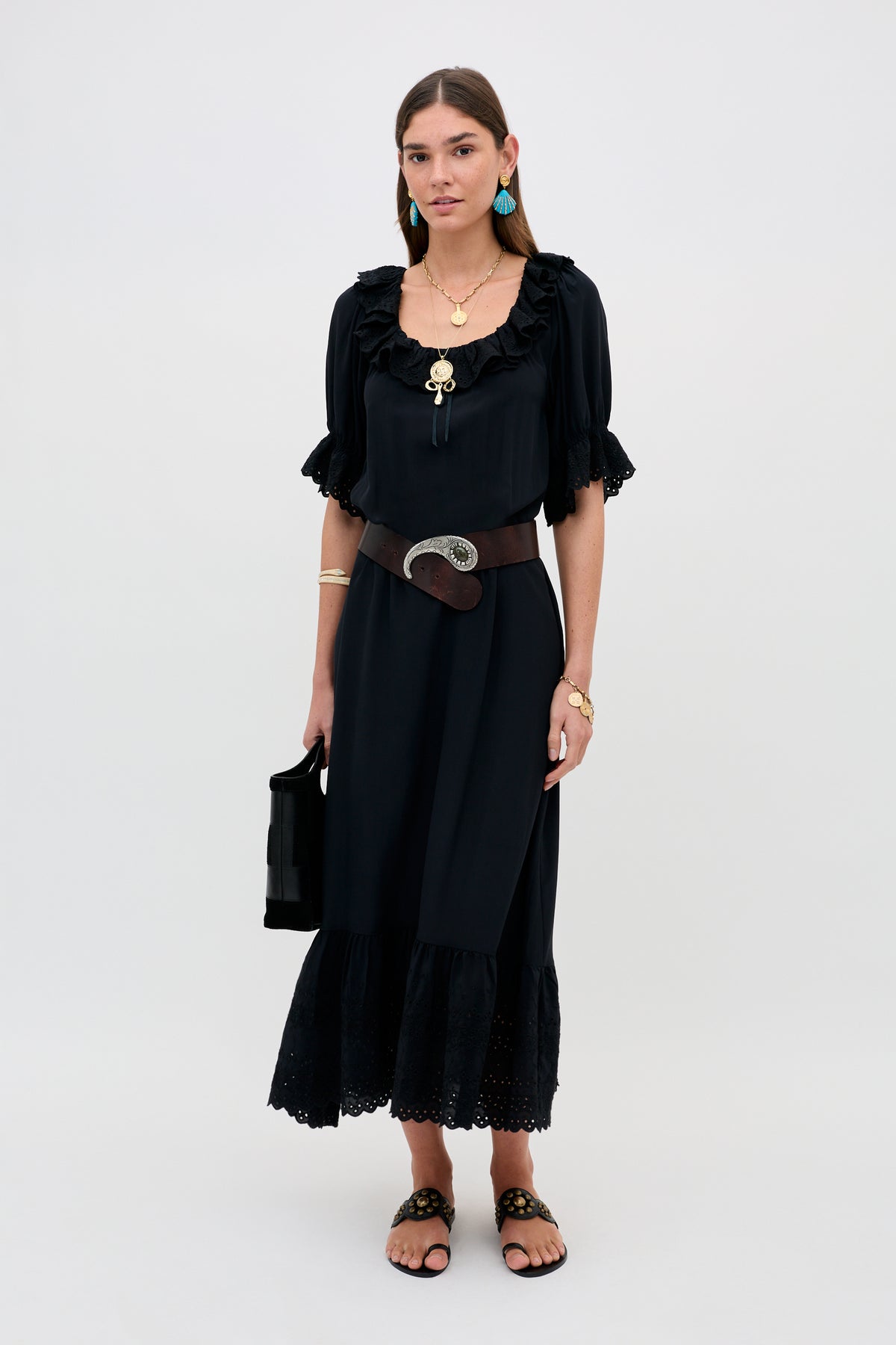A woman poses against a plain background in the Aprilia Gracie Embroidery Black slip dress featuring short sleeves and ruffles, styled with a wide brown belt, layered gold jewelry, blue earrings, black sandals, and a black handbag.