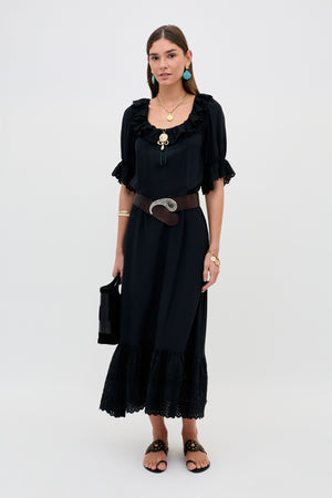 A woman poses against a plain background in the Aprilia Gracie Embroidery Black slip dress featuring short sleeves and ruffles, styled with a wide brown belt, layered gold jewelry, blue earrings, black sandals, and a black handbag.