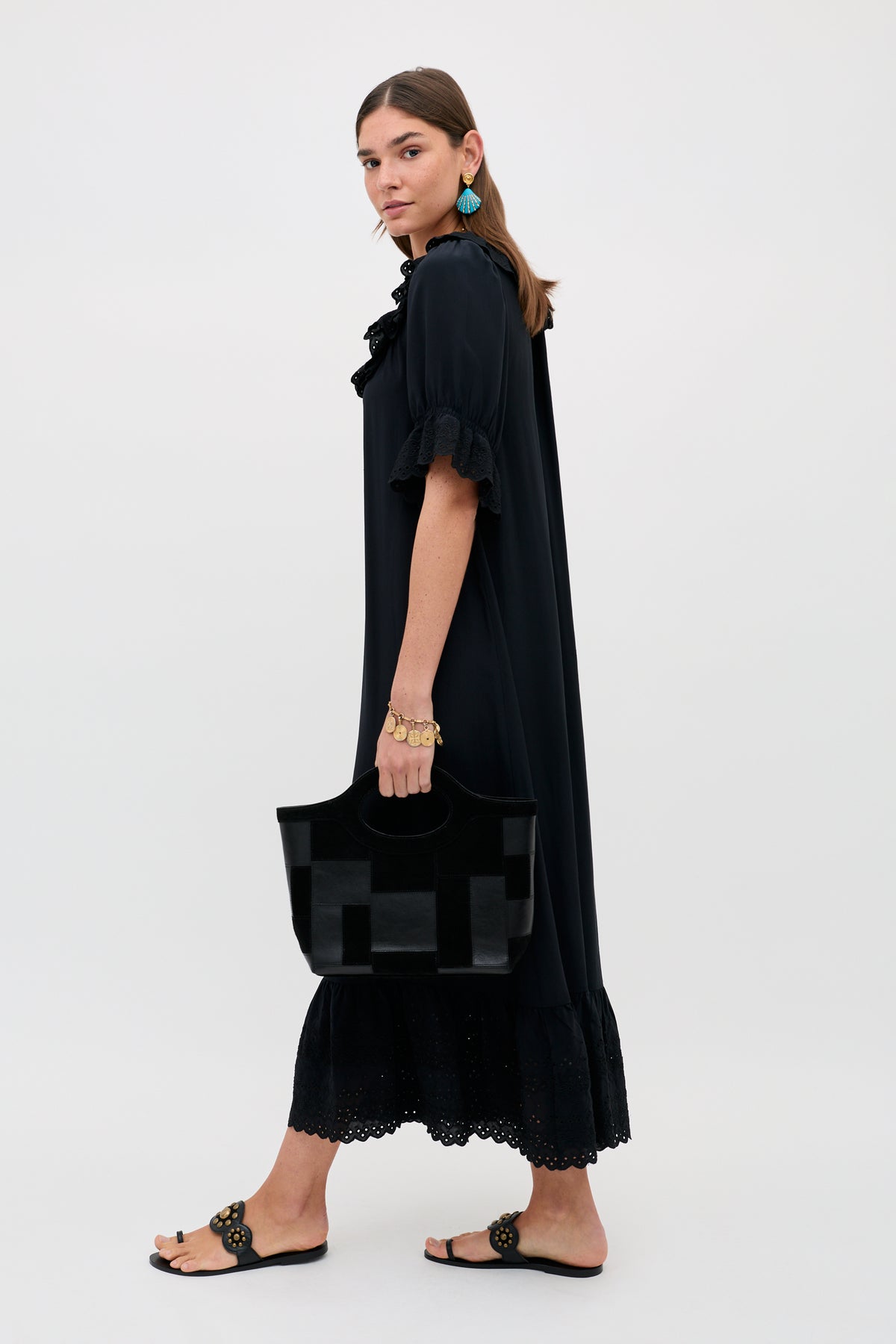 Wearing the Aprilia Gracie Embroidery Black slip dress with lace trim, a woman pairs it with black sandals, blue earrings, gold jewelry, and carries a large black handbag against a plain light background.