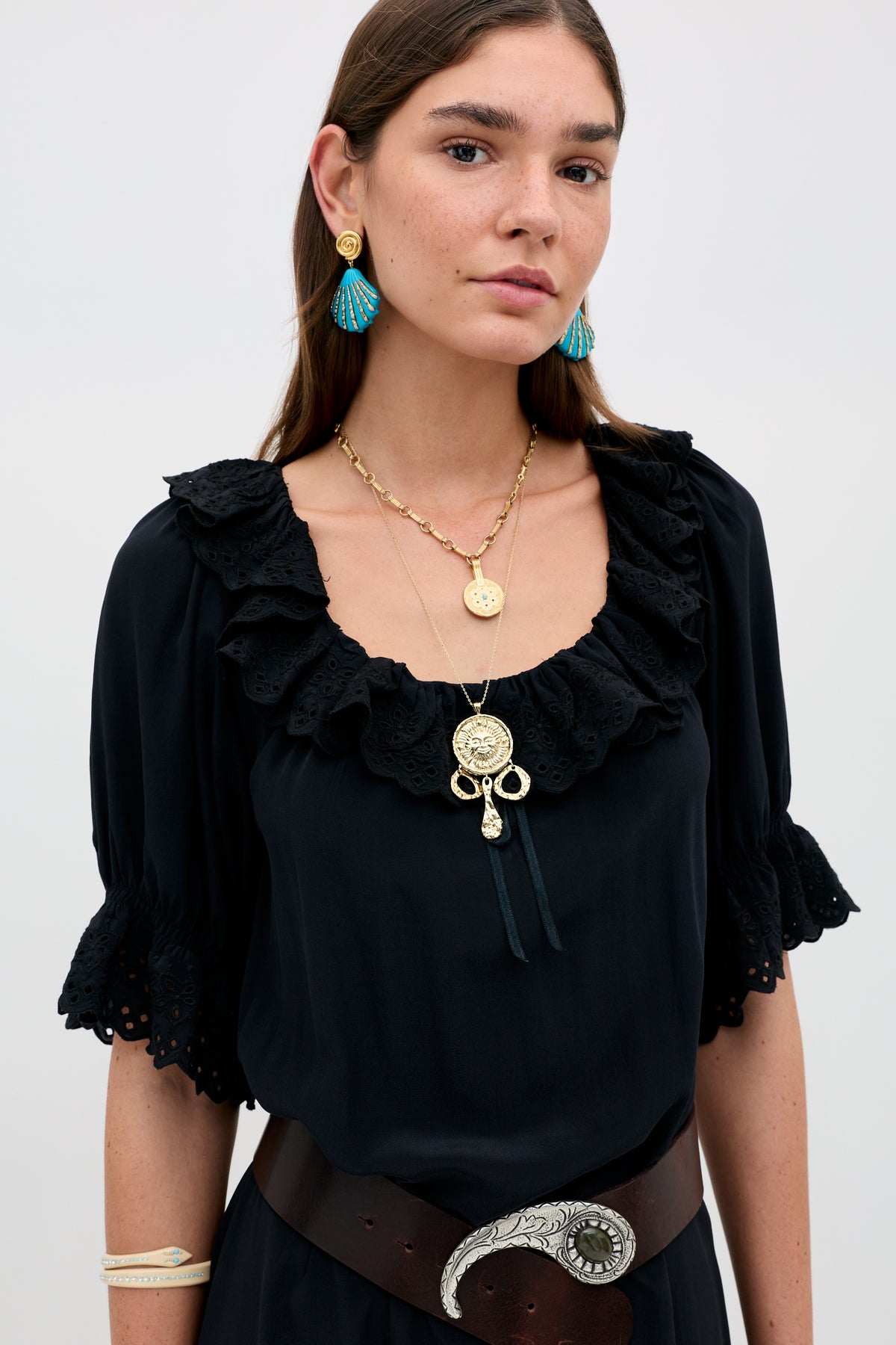 A woman with long brown hair wears the Aprilia Gracie Embroidery Black slip dress with a wide brown belt, gold layered necklaces, and gold and blue shell earrings, standing against a plain light background.