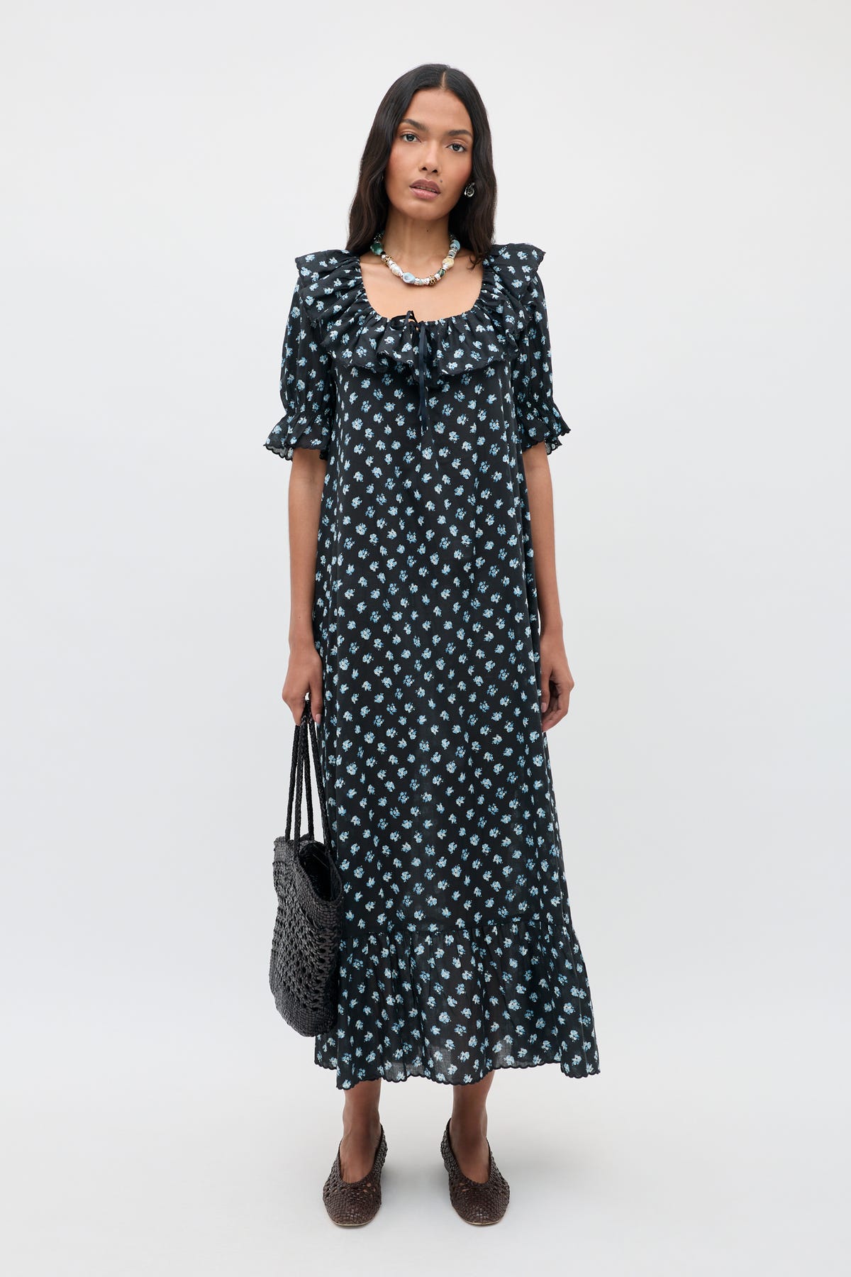 Woman stands against a plain background wearing the Aprilia - Seville Floral Black dress, an ankle-length black dress with white floral patterns, ruffled sleeves, and hem. She carries a black woven bag and wears brown shoes and a necklace.