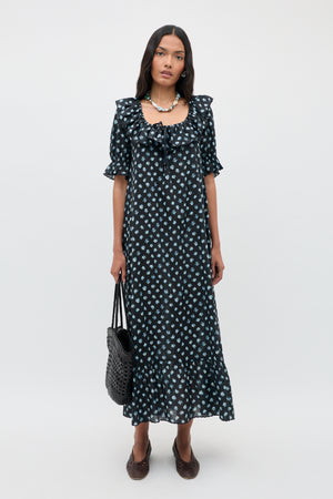 Woman stands against a plain background wearing the Aprilia - Seville Floral Black dress, an ankle-length black dress with white floral patterns, ruffled sleeves, and hem. She carries a black woven bag and wears brown shoes and a necklace.