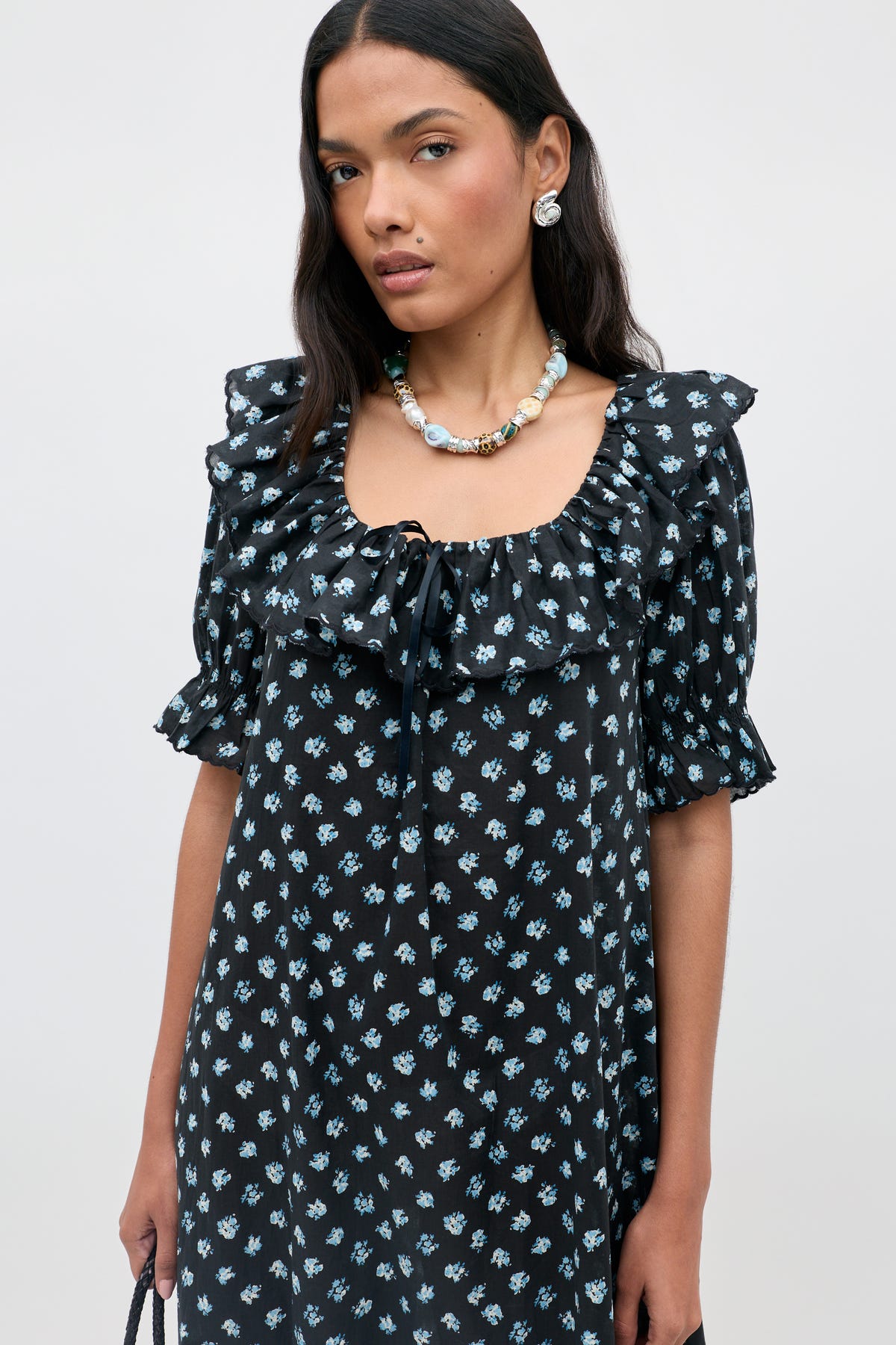 A woman wears the "Aprilia - Seville Floral Black" dress, featuring small blue floral prints, a ruffled neckline, and puff sleeves. She accessorizes with a beaded necklace, pearl earrings, and carries a black bag. The background is plain light gray.