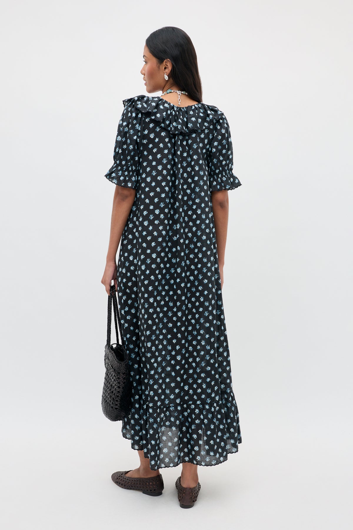 A person stands facing away, wearing the Aprilia - Seville Floral Black dress, an ankle-length black dress adorned with white floral patterns, featuring ruffled sleeves and a ruffled hem. They carry a black woven bag and wear woven shoes. Their hair is long and straight.
