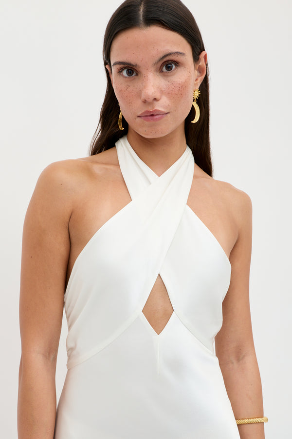 A woman with long brown hair wears the Dune - Satin Gown, a vintage-inspired halterneck dress featuring a crisscross front and small cutout. She accessorizes with gold hoop earrings and a bracelet, standing against a plain light background.