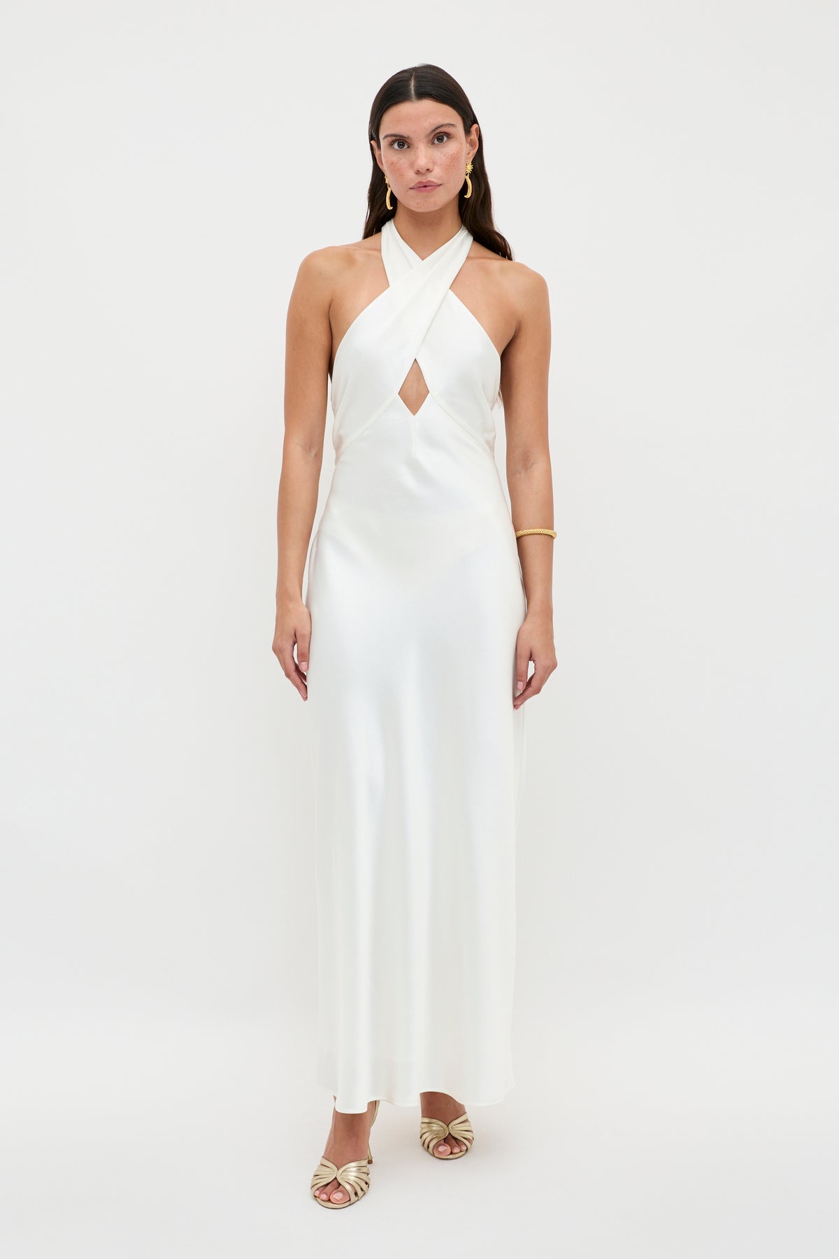 A woman poses against a plain white background wearing the Dune - Satin Gown, a vintage-inspired halterneck dress with a crisscross neckline and front cutout. She has straight brown hair, gold sandals, and a gold bracelet.