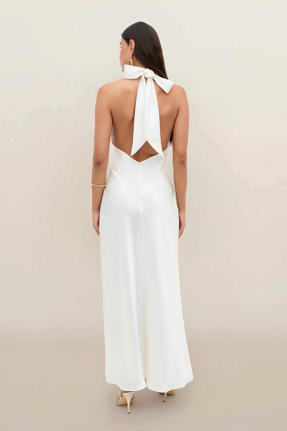 A woman stands facing away, wearing the Dune Satin Gown—an elegant white halterneck dress with an open back and a bow tied behind her neck. She has long brown hair and is accessorized with gold heels and a bracelet. The background is plain beige.