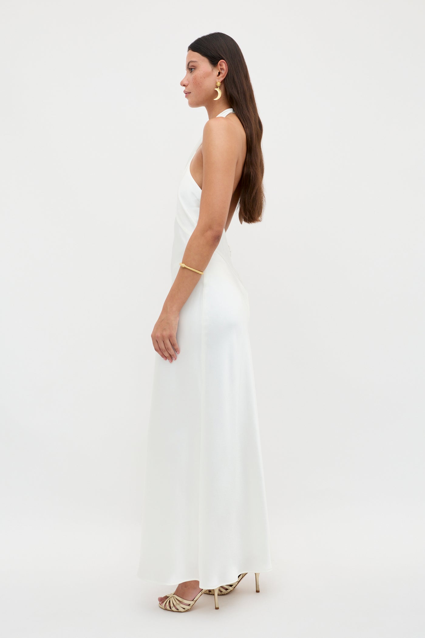A woman with long brown hair, wearing the Dune Satin Gown and gold high-heeled sandals, poses in profile against a plain white background. She accessorizes with gold earrings and a gold bracelet.