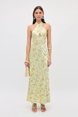 A woman in the Dune - Botanical Embroidery Green halterneck dress holds a gold purse. She has straight brown hair, turquoise earrings, gold bracelets, and leopard-print heeled sandals against a plain white background.