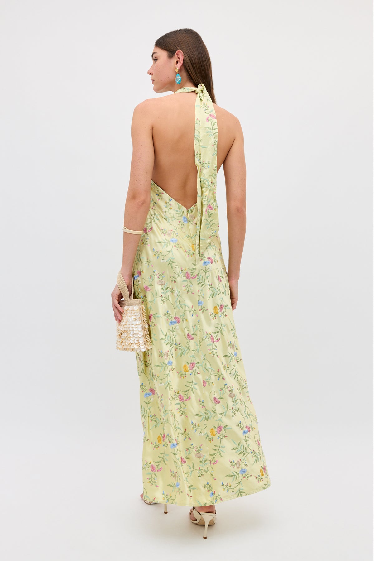 A woman stands facing away in the Dune - Botanical Embroidery Green halterneck floral dress and high heels, holding a beaded handbag. She accessorizes with turquoise earrings and a bracelet against a plain white background.