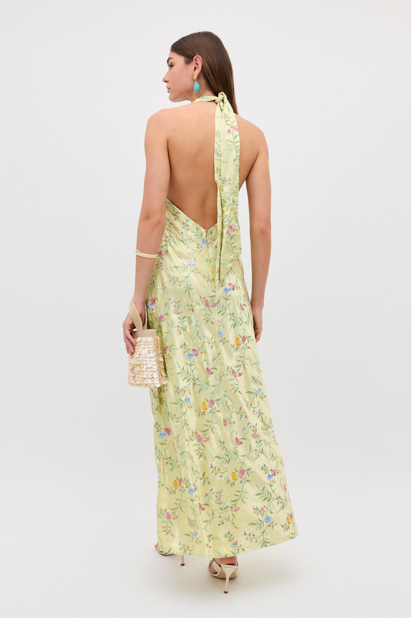 A woman stands facing away in the Dune - Botanical Embroidery Green halterneck floral dress and high heels, holding a beaded handbag. She accessorizes with turquoise earrings and a bracelet against a plain white background.