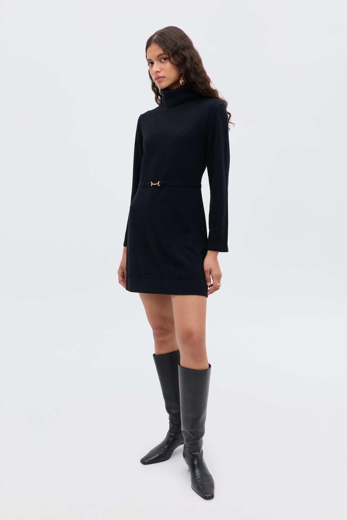 A woman stands confidently in the Aly - Black, a long-sleeve black turtleneck mini dress with a belt detail, paired with knee-high black boots for a chic sixties-inspired look against a plain light background.