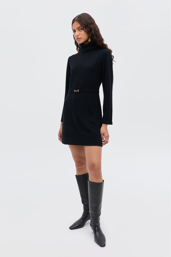 A woman stands confidently in the Aly - Black, a long-sleeve black turtleneck mini dress with a belt detail, paired with knee-high black boots for a chic sixties-inspired look against a plain light background.
