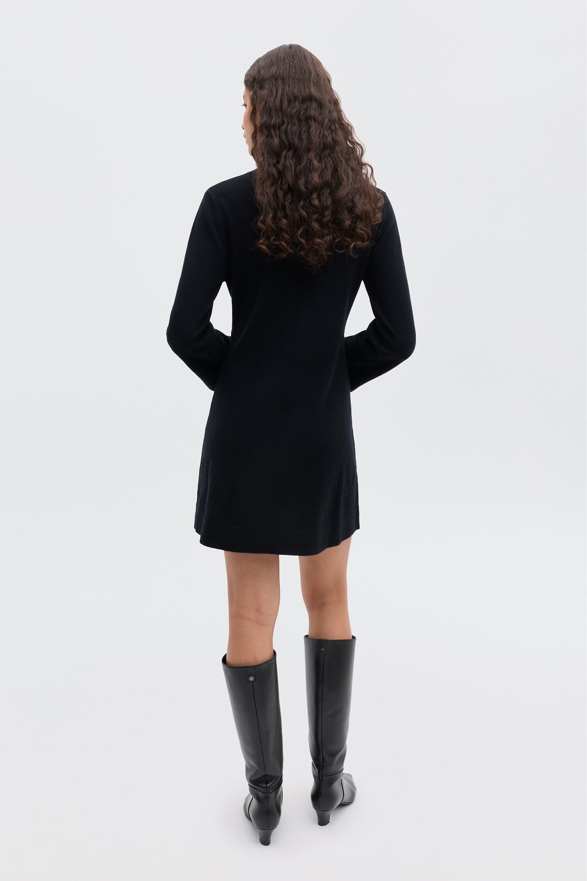 A person with long, curly brown hair stands with their back to the camera, wearing the Aly - Black winter LBD and knee-high black boots against a plain light background.