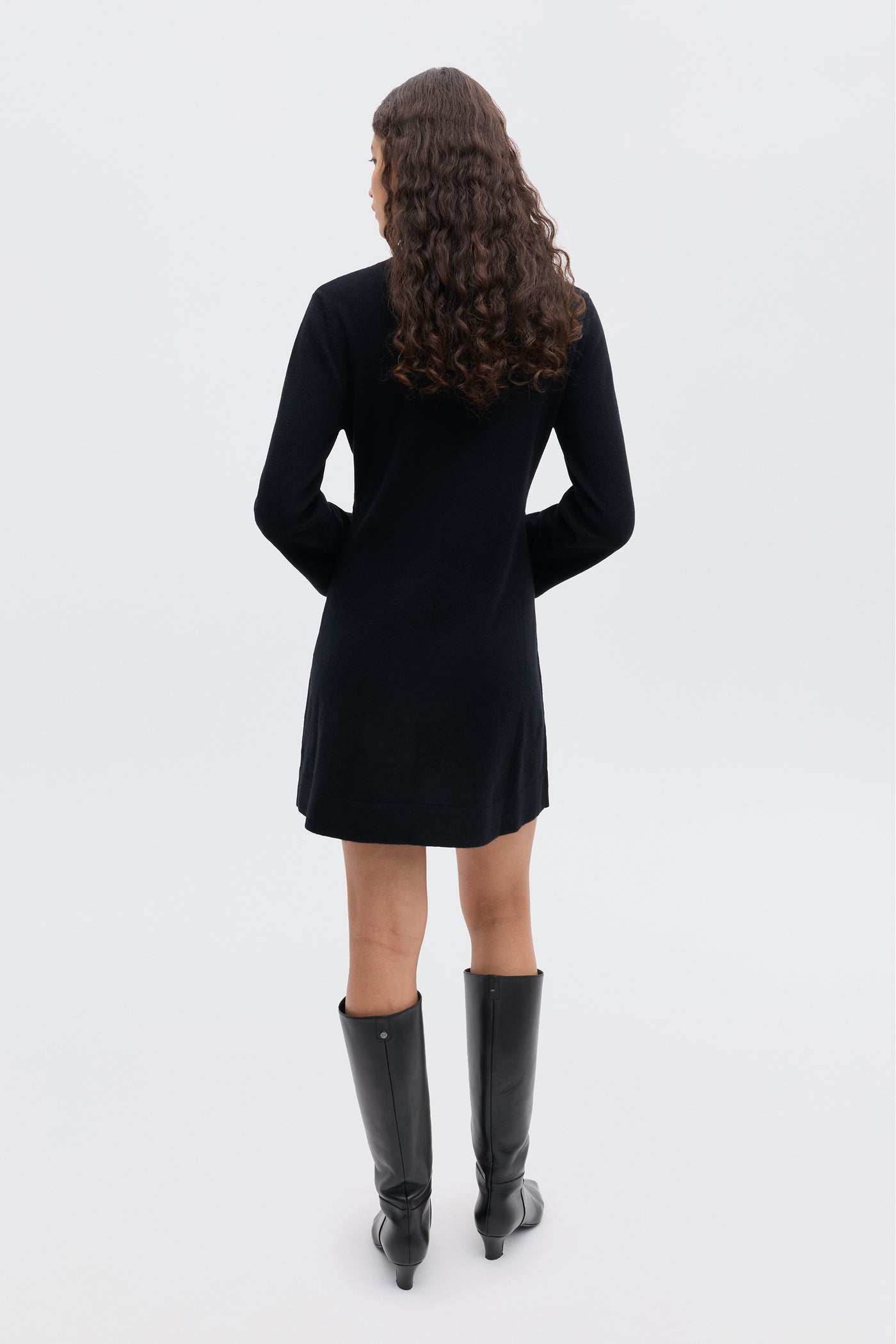 A person with long, curly brown hair stands with their back to the camera, wearing the Aly - Black winter LBD and knee-high black boots against a plain light background.